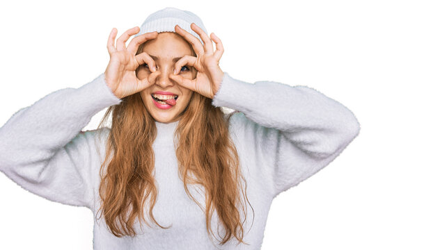 Young caucasian girl wearing wool sweater and winter cap doing ok gesture like binoculars sticking tongue out, eyes looking through fingers. crazy expression.