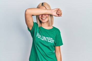 Beautiful blonde woman wearing volunteer t shirt smiling cheerful playing peek a boo with hands showing face. surprised and exited