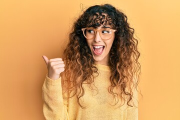 Young hispanic girl wearing casual clothes and glasses pointing thumb up to the side smiling happy with open mouth