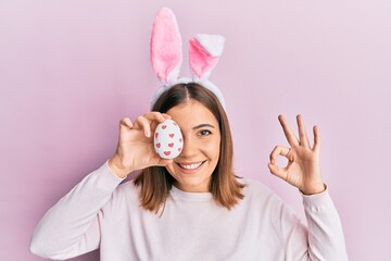 Obraz na płótnie Canvas Young beautiful woman wearing cute easter bunny ears holding egg doing ok sign with fingers, smiling friendly gesturing excellent symbol