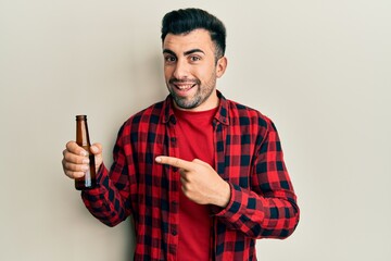 Young hispanic man drinking a bottle of beer smiling happy pointing with hand and finger