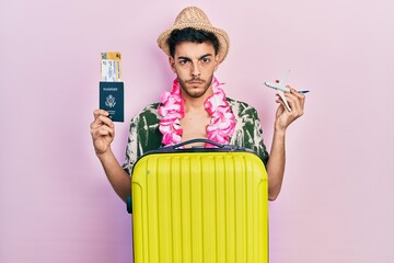 Young hispanic man wearing summer style and hawaiian lei holding passport and plane toy skeptic and...