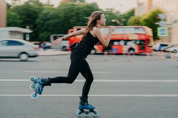 Young fit woman on roller skates with wheels rollerblades during summer day on busy road with transport leads active lifestyle wears black sportsclothes breathes fresh air. Movement concept. - obrazy, fototapety, plakaty