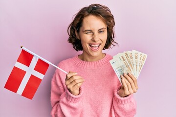 Young brunette woman holding norway flag and krone banknotes winking looking at the camera with...