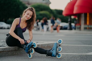 Outdoor shot of young sporty woman in sportswears puts on roller blades poses on road going to ride...