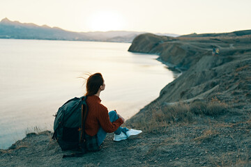 woman hiker sits on the ground in the mountains near the sea at sunset
