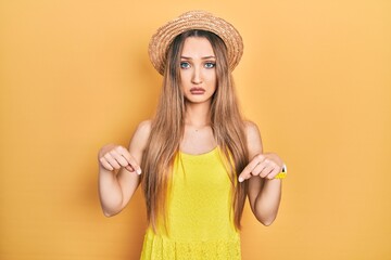 Young blonde girl wearing summer hat pointing down looking sad and upset, indicating direction with...