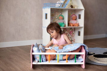 Cute little girl is playing with a doll at home. The child puts the doll to sleep in a toy bed....