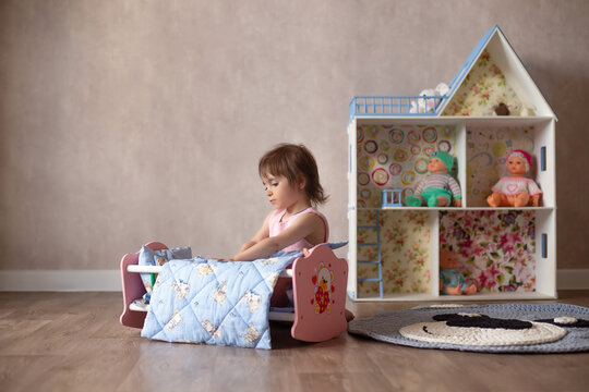 Cute little girl is playing with a doll at home. The child puts the doll to sleep in a toy bed. Role-playing games for children.