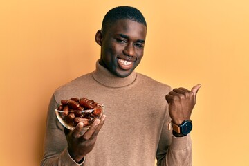 Young african american man holding bowl with dates pointing thumb up to the side smiling happy with open mouth