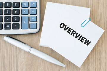 a stack of paper for notes, on one text OVERVIEW on a wooden background next to a calculator, silver pen