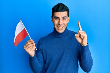 Handsome hispanic man holding poland flag smiling with an idea or question pointing finger with happy face, number one