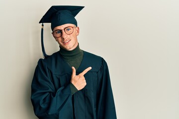 Young caucasian man wearing graduation cap and ceremony robe cheerful with a smile of face pointing with hand and finger up to the side with happy and natural expression on face