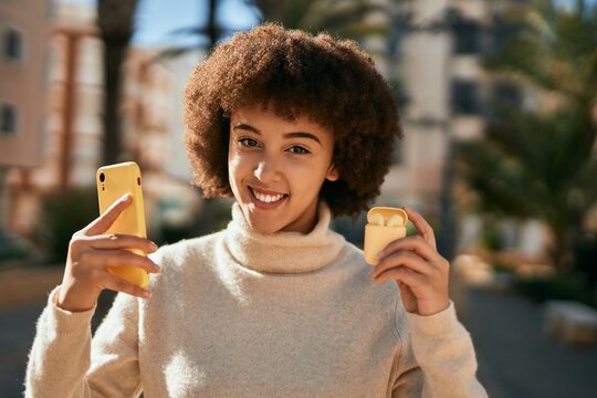 Young hispanic girl smiling happy using smartphone and holding earphones at the city.