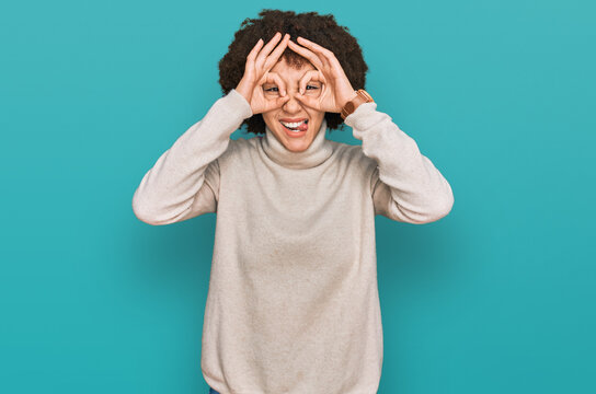 Young hispanic girl wearing wool winter sweater doing ok gesture like binoculars sticking tongue out, eyes looking through fingers. crazy expression.