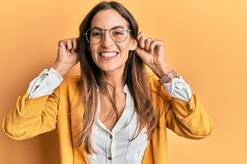 Young beautiful woman wearing business style and glasses smiling pulling ears with fingers, funny...