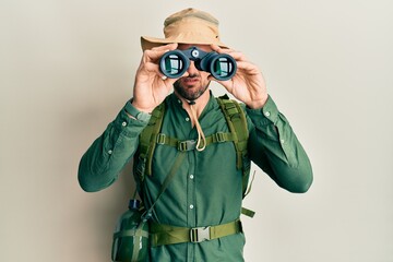 Handsome man with beard wearing explorer hat looking through binoculars clueless and confused expression. doubt concept. - obrazy, fototapety, plakaty
