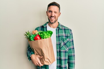 Handsome man with beard holding paper bag with groceries winking looking at the camera with sexy...