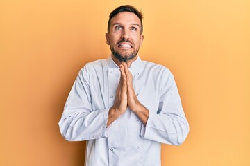 Handsome man with beard wearing professional cook uniform begging and praying with hands together with hope expression on face very emotional and worried. begging.