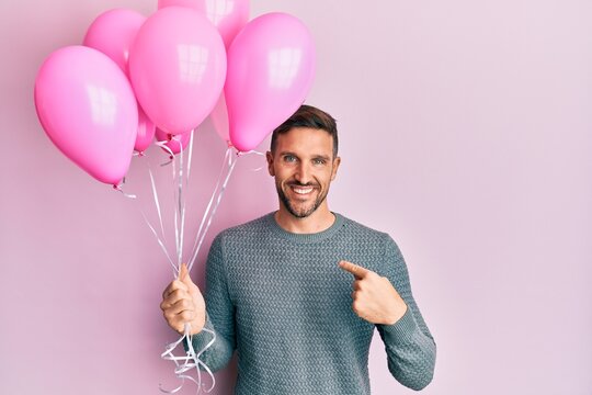Handsome man with beard holding pink balloons pointing finger to one self smiling happy and proud