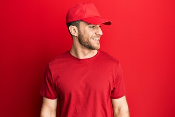 Young caucasian man wearing delivery uniform and cap looking away to side with smile on face, natural expression. laughing confident.
