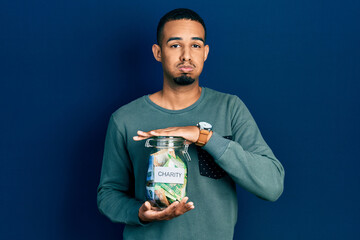 Young african american man holding charity jar with south africa rand banknotes puffing cheeks with funny face. mouth inflated with air, catching air.