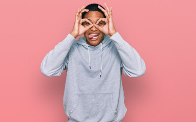 Young handsome hispanic man wearing casual sweatshirt doing ok gesture like binoculars sticking tongue out, eyes looking through fingers. crazy expression.