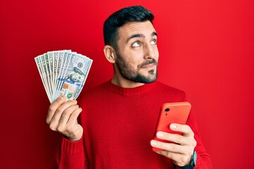 Young hispanic man using smartphone holding usa dollars banknotes smiling looking to the side and staring away thinking.