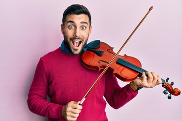 Young hispanic musician man playing violin celebrating crazy and amazed for success with open eyes...