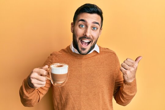 Young hispanic man holding coffee pointing thumb up to the side smiling happy with open mouth