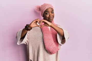 Young african american woman wearing wool hat and winter scarf smiling in love doing heart symbol...