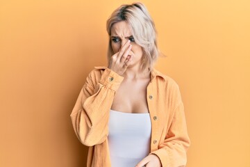 Young blonde girl wearing casual clothes smelling something stinky and disgusting, intolerable smell, holding breath with fingers on nose. bad smell - obrazy, fototapety, plakaty