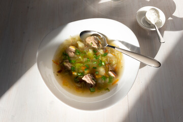 Soup with meat, chanterelle mushrooms, vegetables in white bowl, sour cream, vase with lilac on white table in sun light