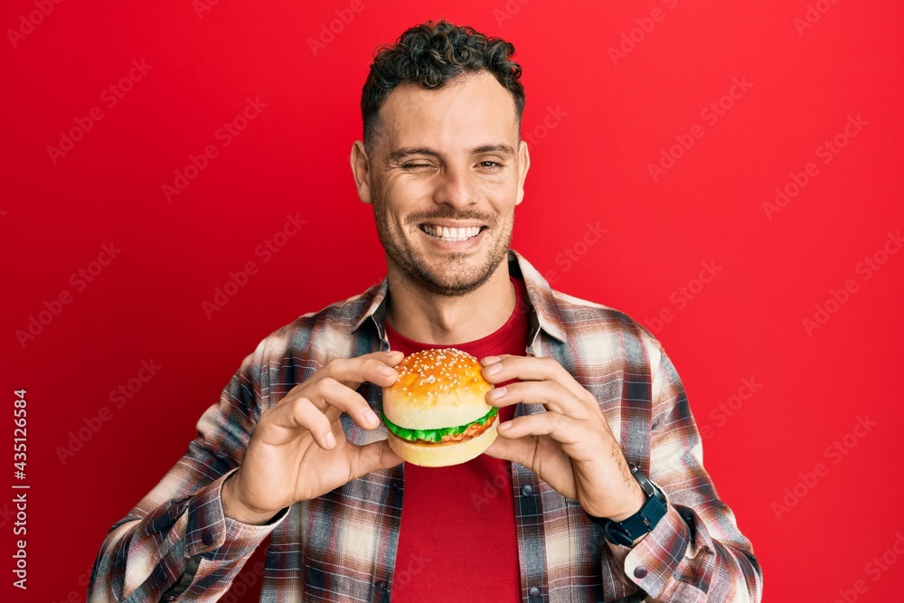 Wall mural young hispanic man eating a tasty classic burger winking looking at the camera with sexy expression,