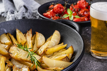 Roasted potatoes with rosemary in iron casserole and plate of confit tomatoes on dark background