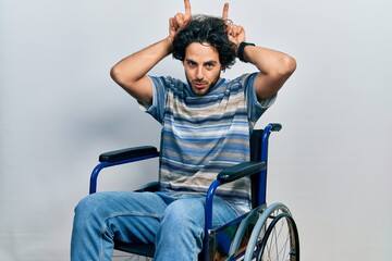 Obraz premium Handsome hispanic man sitting on wheelchair doing funny gesture with finger over head as bull horns