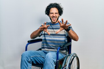 Handsome hispanic man sitting on wheelchair smiling funny doing claw gesture as cat, aggressive and...