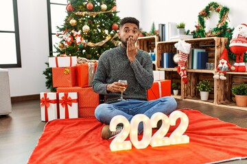Obraz na płótnie Canvas Young hispanic man with beard sitting by christmas tree celebrating 2022 new year covering mouth with hand, shocked and afraid for mistake. surprised expression
