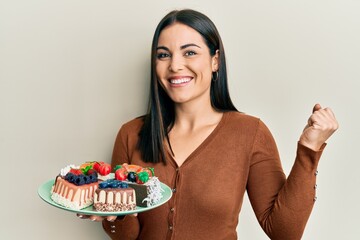 Young brunette woman holding plate with cake slices screaming proud, celebrating victory and success very excited with raised arm