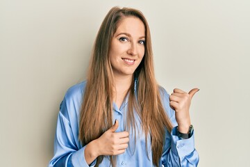 Young blonde woman wearing casual blue shirt smiling with happy face looking and pointing to the side with thumb up.