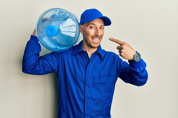 Bald courier man with beard holding a gallon bottle of water for delivery smiling happy pointing with hand and finger
