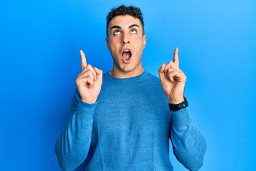 Hispanic young man wearing casual winter sweater amazed and surprised looking up and pointing with fingers and raised arms.