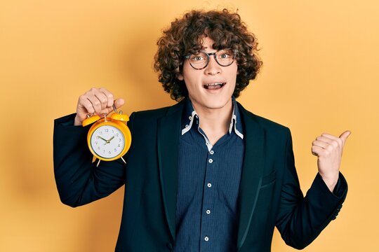 Handsome young man wearing business jacket and holding clock pointing thumb up to the side smiling happy with open mouth