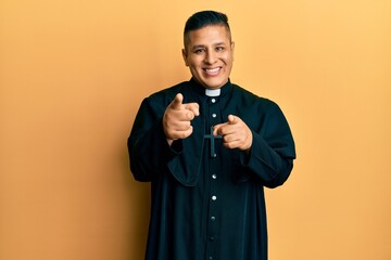 Young latin priest man standing over yellow background pointing fingers to camera with happy and...