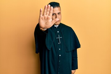 Young latin priest man standing over yellow background doing stop sing with palm of the hand....