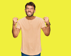 Handsome young man with beard wearing casual tshirt angry and mad raising fists frustrated and furious while shouting with anger. rage and aggressive concept.