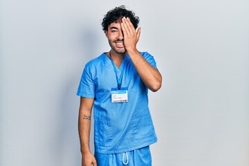 Young hispanic man wearing blue male nurse uniform covering one eye with hand, confident smile on face and surprise emotion.