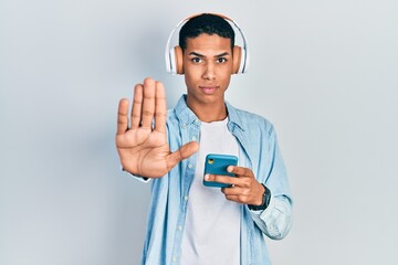 Young african american guy using smartphone wearing headphones with open hand doing stop sign with serious and confident expression, defense gesture