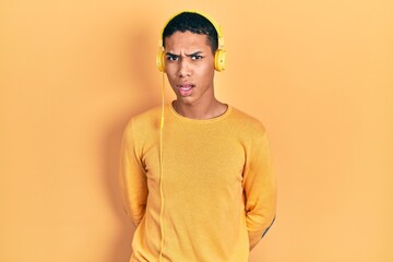Young african american guy listening to music using headphones in shock face, looking skeptical and...