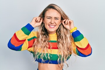 Beautiful young blonde woman wearing colored sweater smiling pulling ears with fingers, funny...